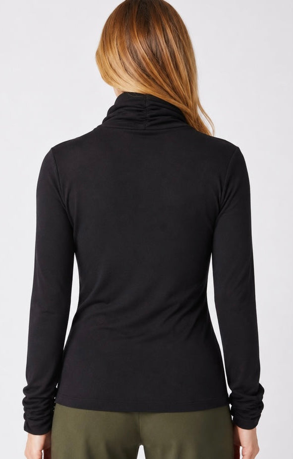 A woman viewed from the back wearing a black long-sleeve cowl-neck top and olive green pants against a light gray studio background.