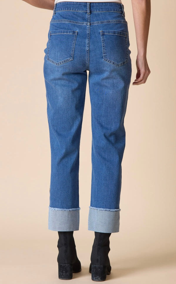 Back view of a person wearing medium-wash blue denim jeans with rolled cuffs and black ankle boots, standing against a plain beige studio background.