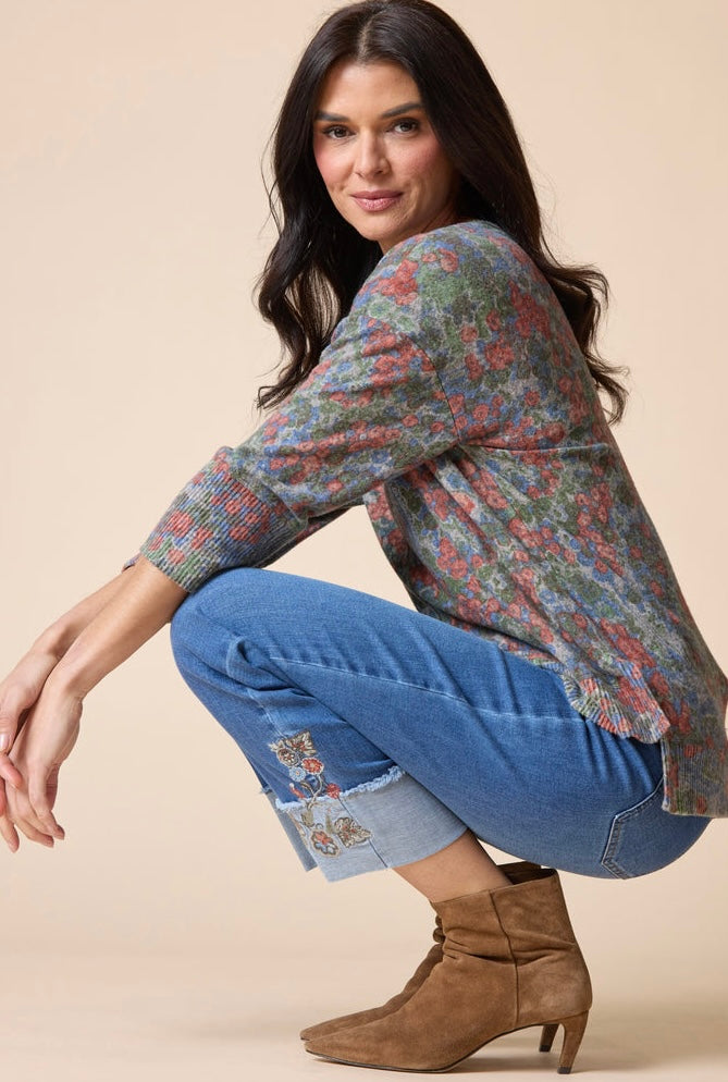 A woman crouching in a studio setting, wearing a long-sleeve knit top with a muted floral pattern in blue, green, and red tones, paired with blue jeans and brown ankle boots against a solid beige background.