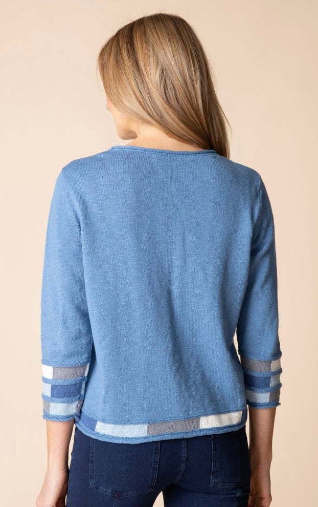 A woman viewed from the back wearing a heathered blue knit sweater with striped trim at the cuffs and hem, paired with dark blue jeans against a solid beige background.