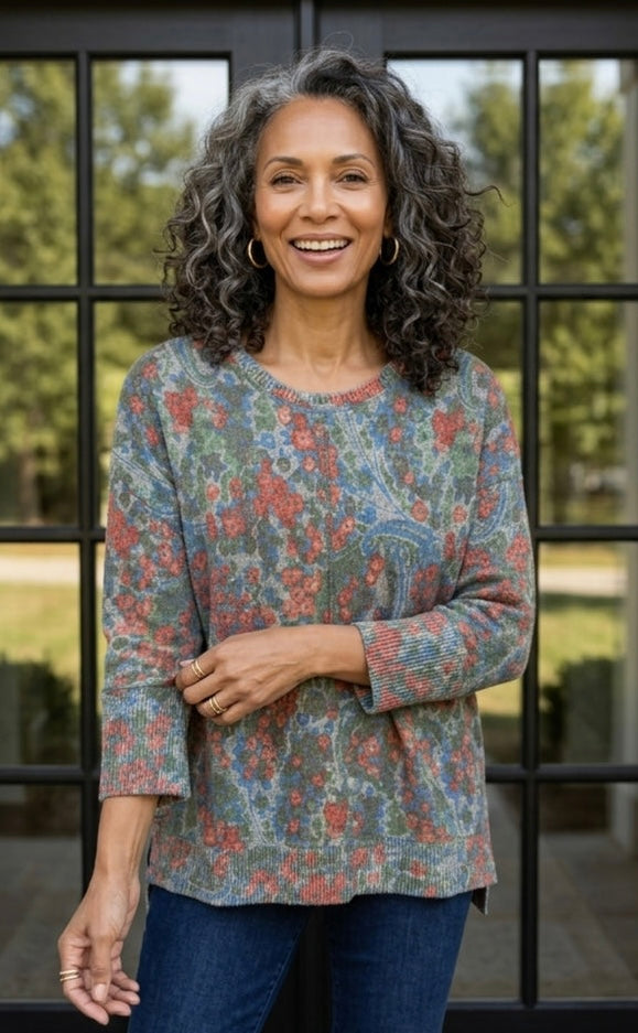 A smiling woman with curly gray hair stands in front of a black-framed glass window, wearing a long-sleeve crewneck top with a blue, green, and red paisley pattern, paired with dark blue jeans.