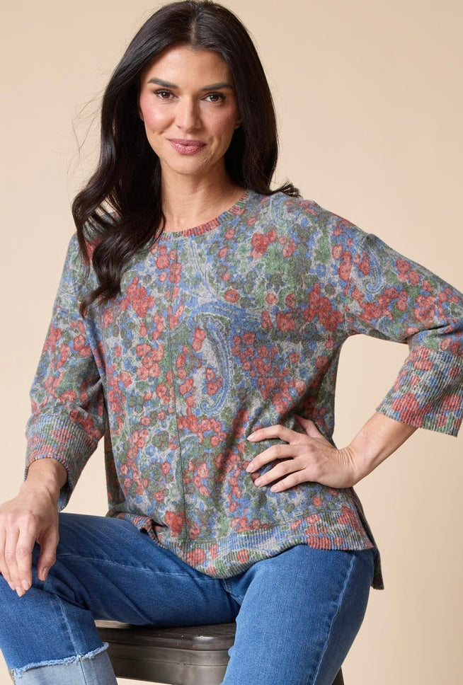 A woman wearing a loose-fitting, three-quarter sleeve knit top with a multicolor paisley pattern in red, blue, green, and beige, paired with blue jeans, seated against a solid beige background.