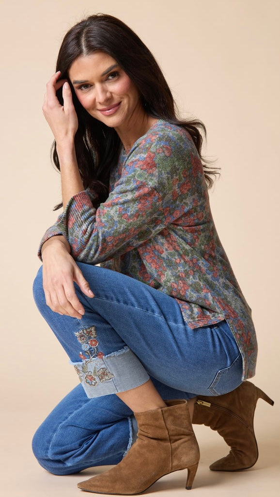 A woman crouching in a studio setting, wearing a long-sleeve floral knit top, blue jeans with rolled cuffs and embroidery, and brown suede ankle boots against a solid beige background.