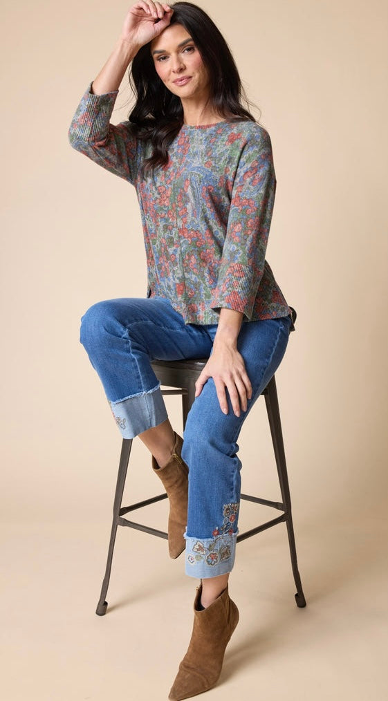A woman sits on a black metal stool wearing a long-sleeve crewneck top with a blue, red, and green floral pattern, blue jeans with cuffed hems, and brown ankle boots against a solid beige background.