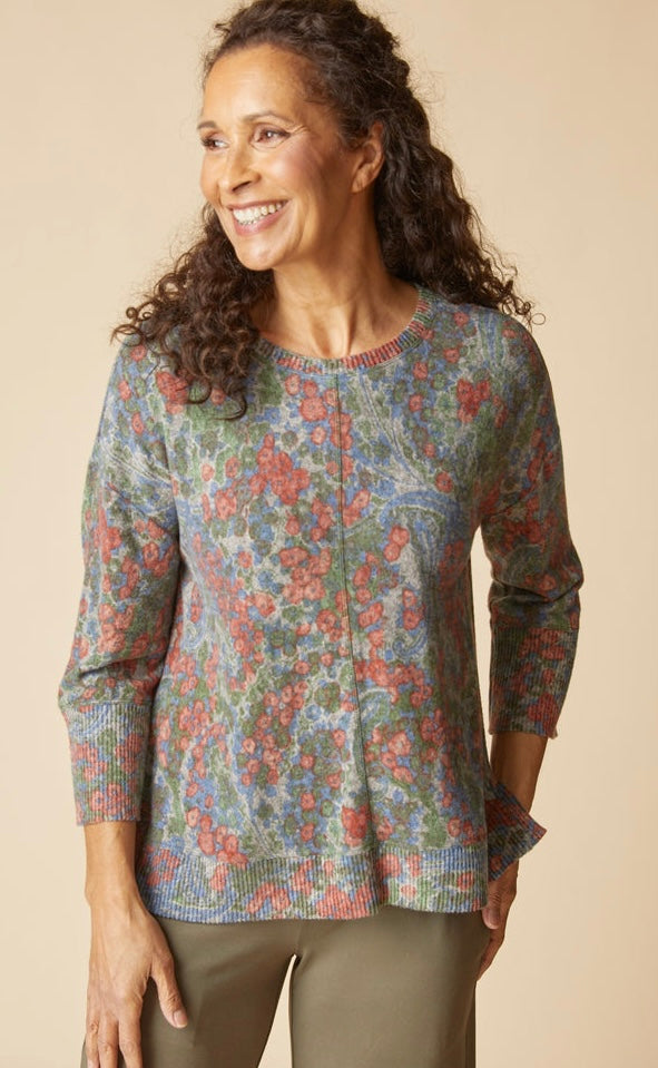 A smiling woman with curly brown hair wearing a long-sleeve knit top with a dense floral pattern in muted green, blue, and coral-red tones, paired with olive pants, against a solid beige studio background.