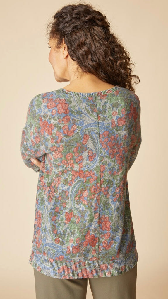 A woman shown from the back wearing a long-sleeve top with a dense paisley and floral pattern in muted blue, green, and coral-red tones, paired with olive pants against a plain beige studio background.
