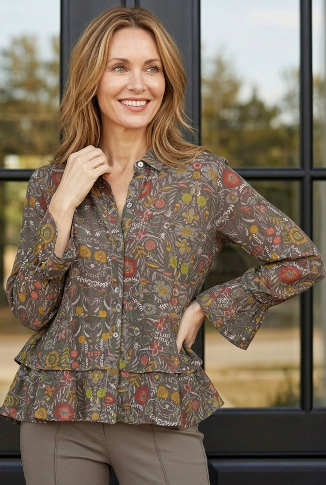 A woman wearing a brown long-sleeve button-up blouse with a colorful abstract floral and paisley print, paired with taupe pants, standing in front of a black-framed window.