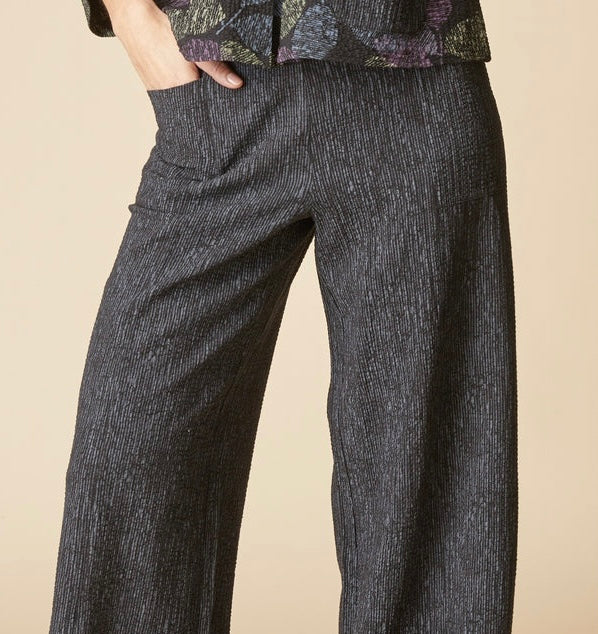 A close-up of a person wearing dark charcoal gray wide-leg textured trousers with a side pocket, paired with a patterned top against a solid beige background.