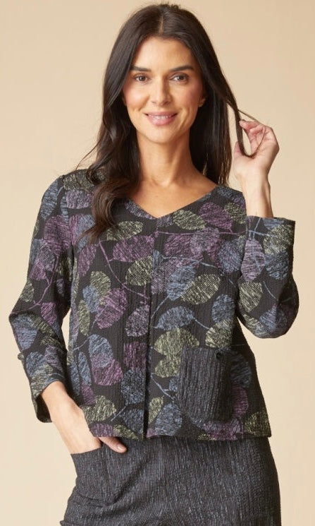A woman modeling a cropped long-sleeve top with a dark abstract leaf pattern in purple, olive green, and black, paired with a textured dark skirt against a solid beige background.