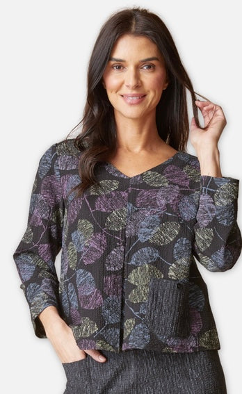 A woman modeling a cropped long-sleeve blouse with a dark base and a leaf-like abstract pattern in purple, olive green, and gray, against a light gray studio background.