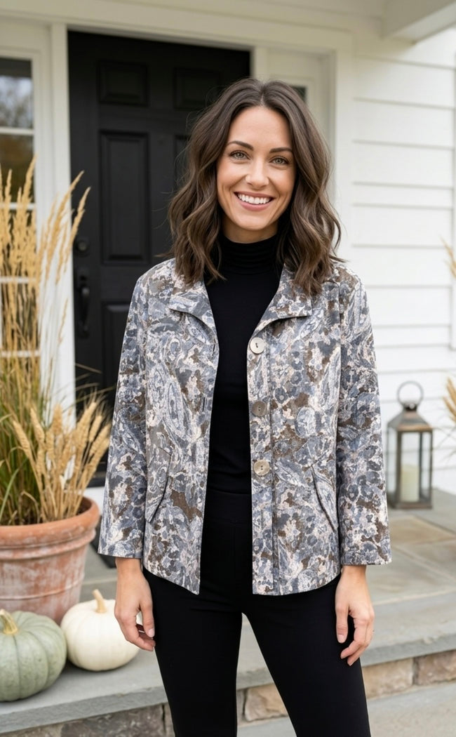 A woman stands on a front porch wearing a long-sleeve button-front jacket with a gray, white, and brown paisley pattern over a black turtleneck and black pants.