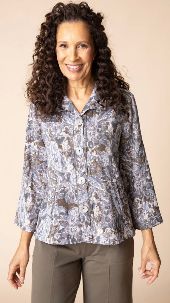 A smiling woman with long curly brown hair wearing a patterned button-up blouse with three-quarter sleeves and brown pants, standing against a solid beige studio background.