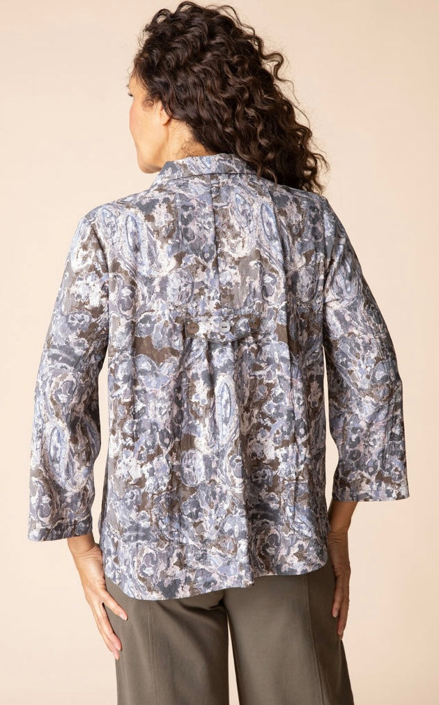 A woman shown from the back wearing a long-sleeve button-up blouse with a gray, white, and brown paisley pattern, paired with olive green pants, against a solid beige background.