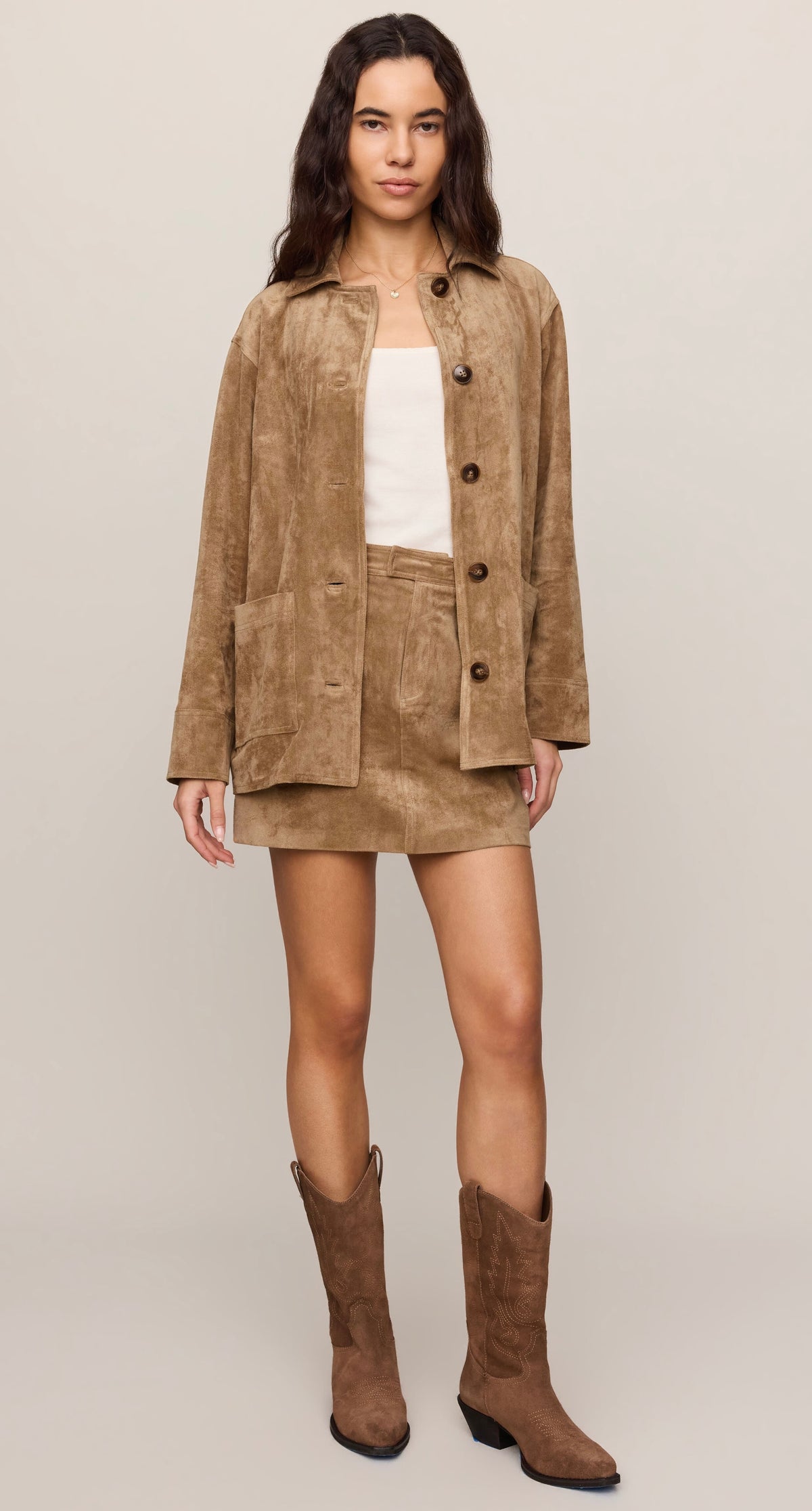 A woman modeling a matching tan suede-like jacket and mini skirt set with a white top and brown cowboy boots, photographed against a plain light beige studio background. -OLIVIA'S