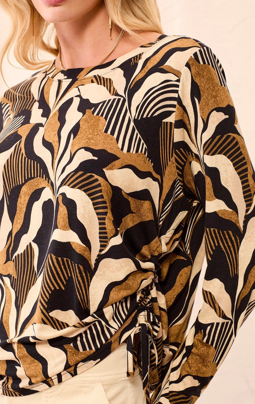 A long-sleeve knit top with a bold abstract botanical pattern in black, tan, and cream, held up on a hanger by a person in a clothing store.