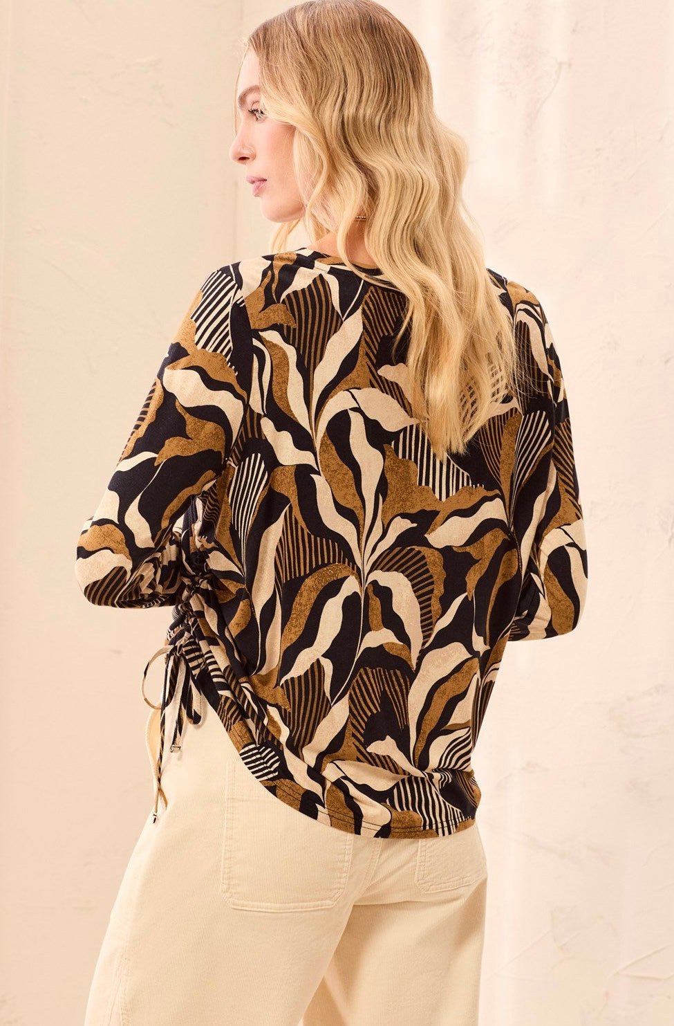 A long-sleeve knit top with a bold abstract botanical pattern in black, tan, and cream, held up on a hanger by a person in a clothing store.