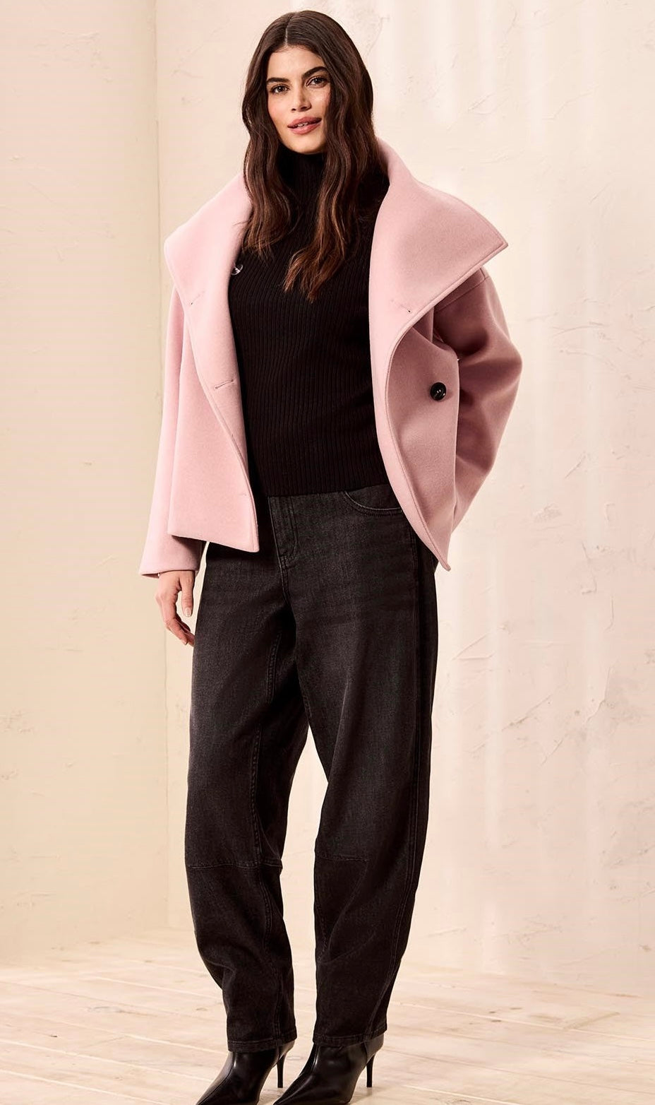 A woman modeling a light pink oversized wrap coat over a black turtleneck sweater, black wide-leg jeans, and black ankle boots, standing against a neutral beige textured wall. - OLIVIA'S