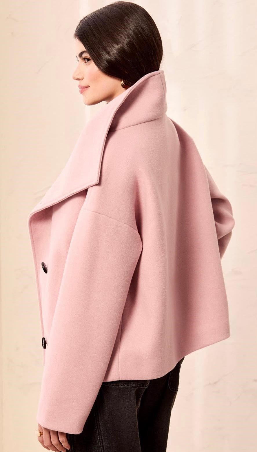 A woman shown from the side wearing a light pink oversized coat with a wide collar and black pants, photographed against a plain light beige background. - OLIVIA'S