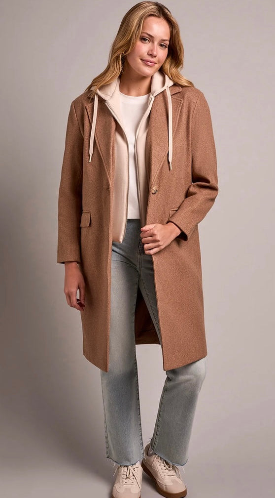A woman modeling a long camel brown hooded coat over a cream hoodie, light blue jeans, and beige sneakers, photographed against a neutral gray studio background. - OLIVIA'S