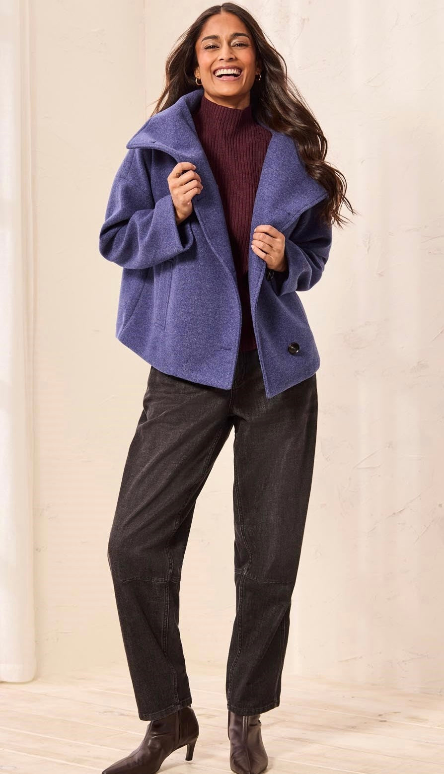 A woman modeling a heathered periwinkle blue open-front coat over a burgundy ribbed turtleneck sweater, paired with dark charcoal pants and brown ankle boots, set against a warm cream-colored background. - OLIVIA'S
