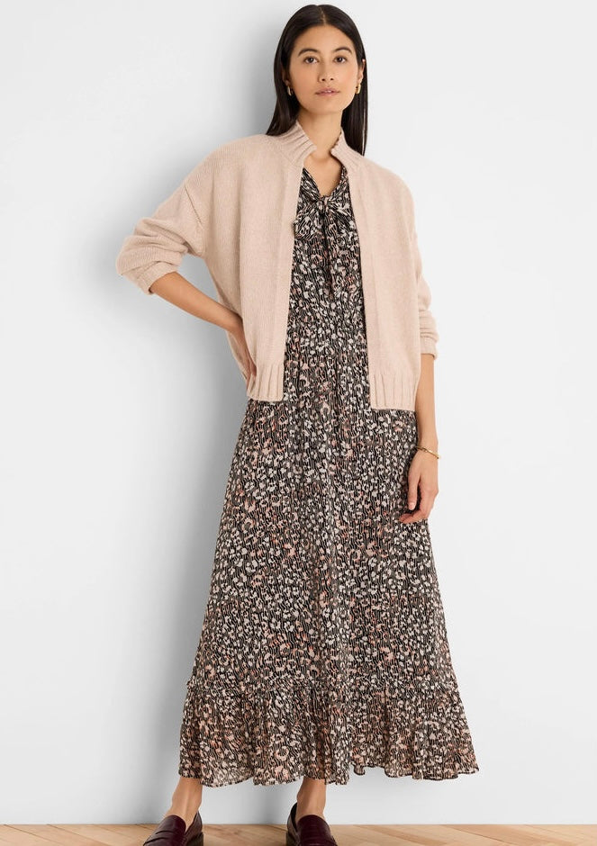 A woman models a light pink knit zip-front cardigan layered over a brown and beige floral print maxi dress, standing against a plain off-white wall.