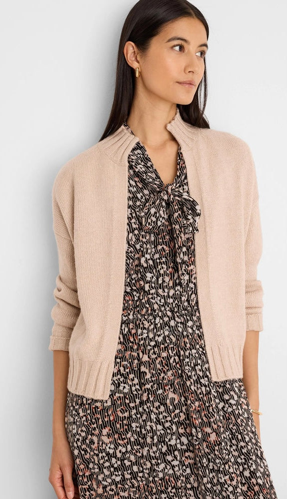 A woman wearing a light beige knit cardigan over a black and white floral print dress, photographed against a clean light gray studio background.