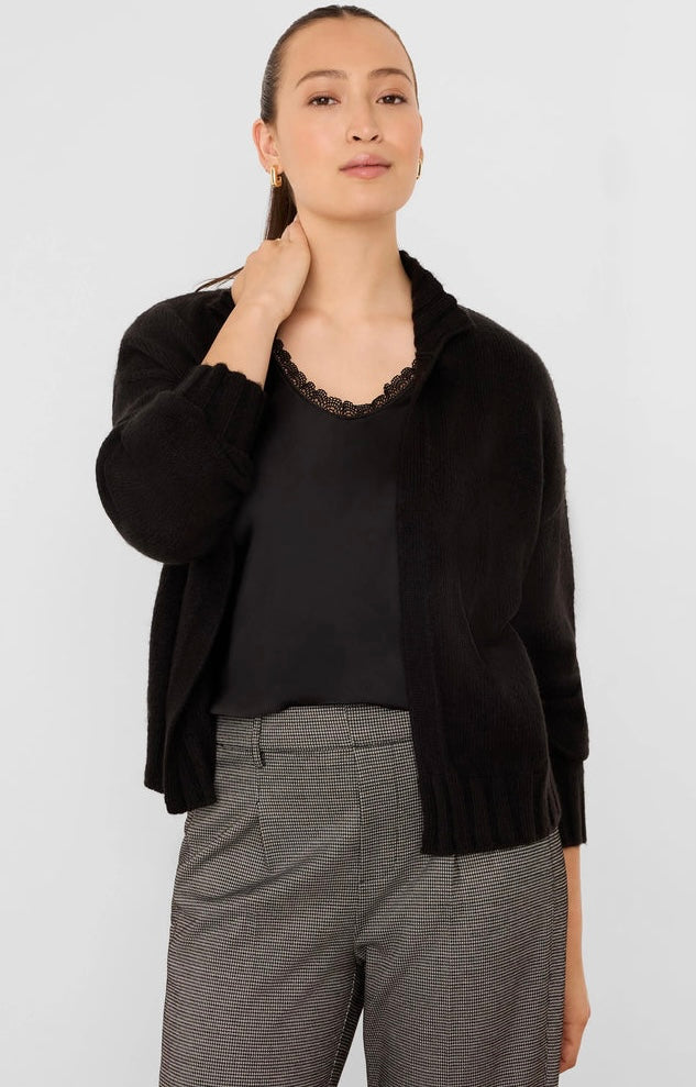 A woman modeling a black knit cardigan with a wide collar over a black top and gray patterned trousers, photographed against a light gray studio background.