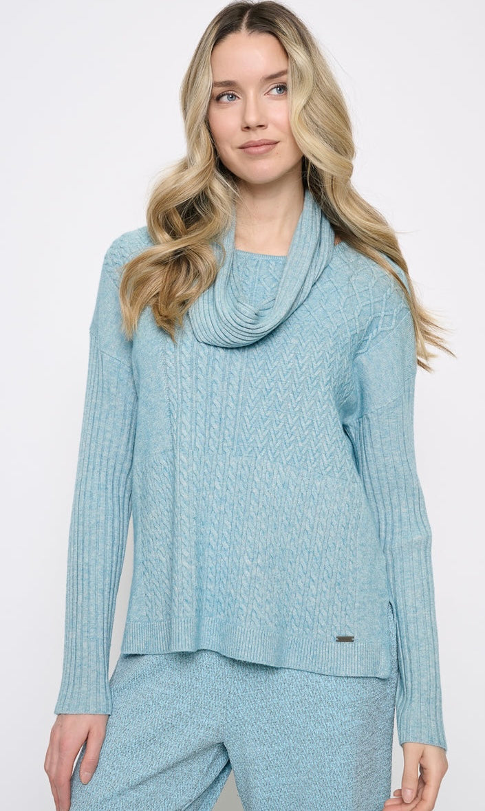 A woman wearing a light blue cable-knit cowl-neck sweater and matching knit pants against a plain white studio background. - OLIVIA'S