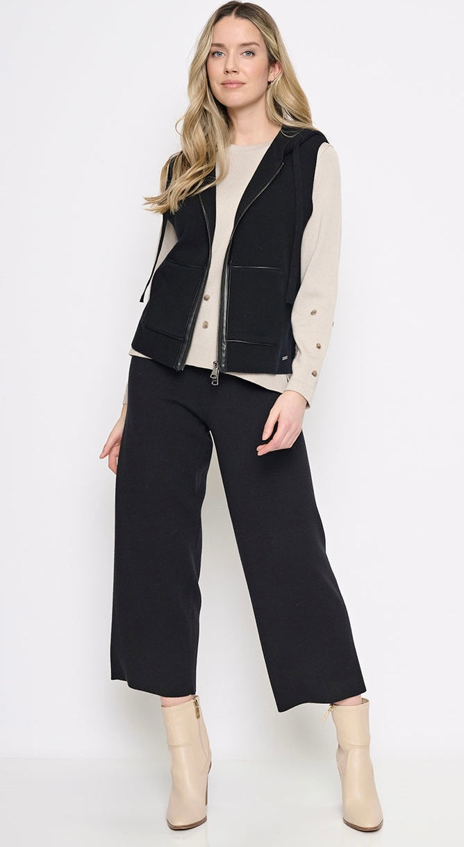 A woman modeling a black sleeveless zip-front vest over a cream long-sleeve top, paired with black wide-leg cropped pants and beige ankle boots, against a plain white studio background. - OLIVIA'S