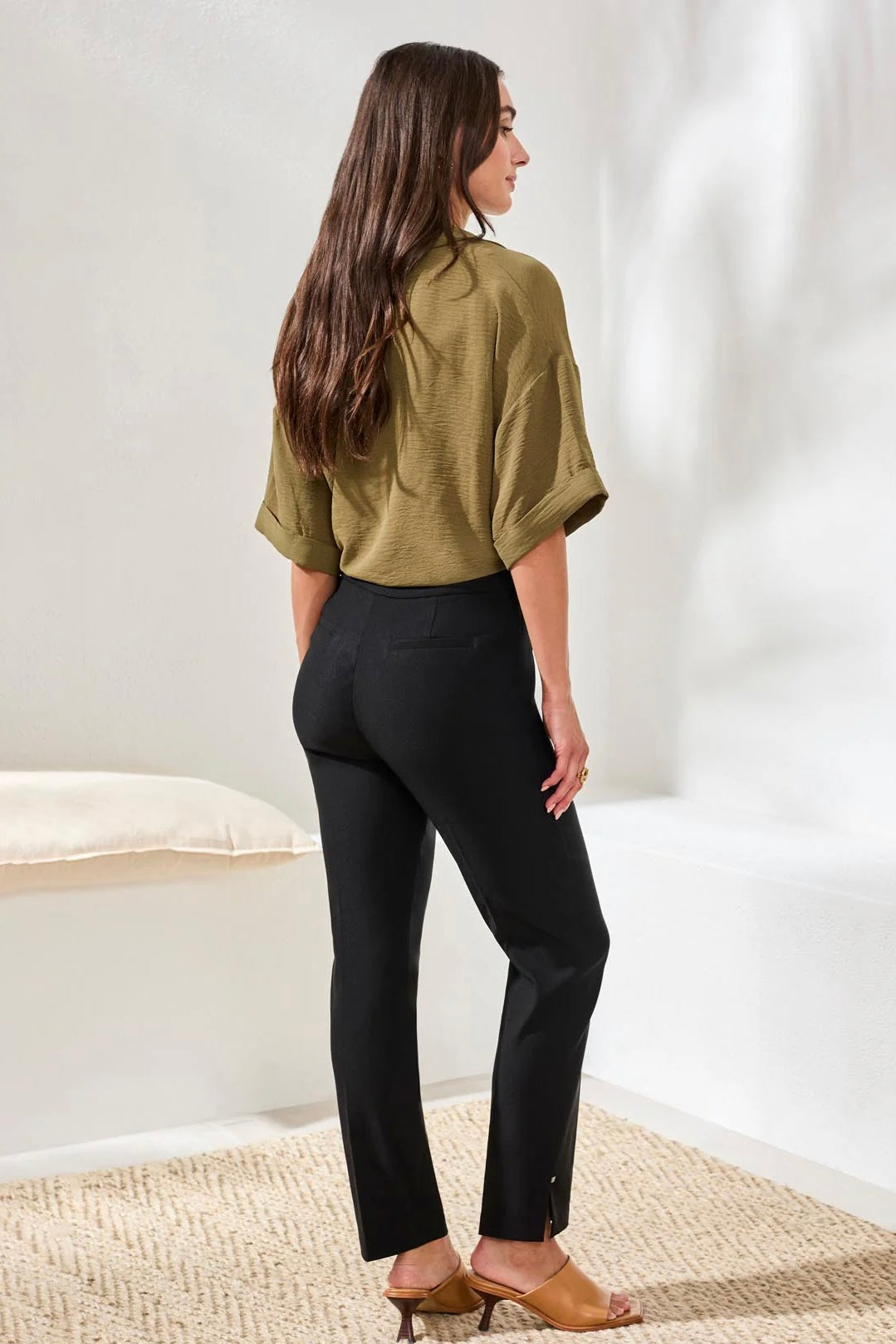 Woman wearing a green blouse and black pants standing in a minimalistic room.