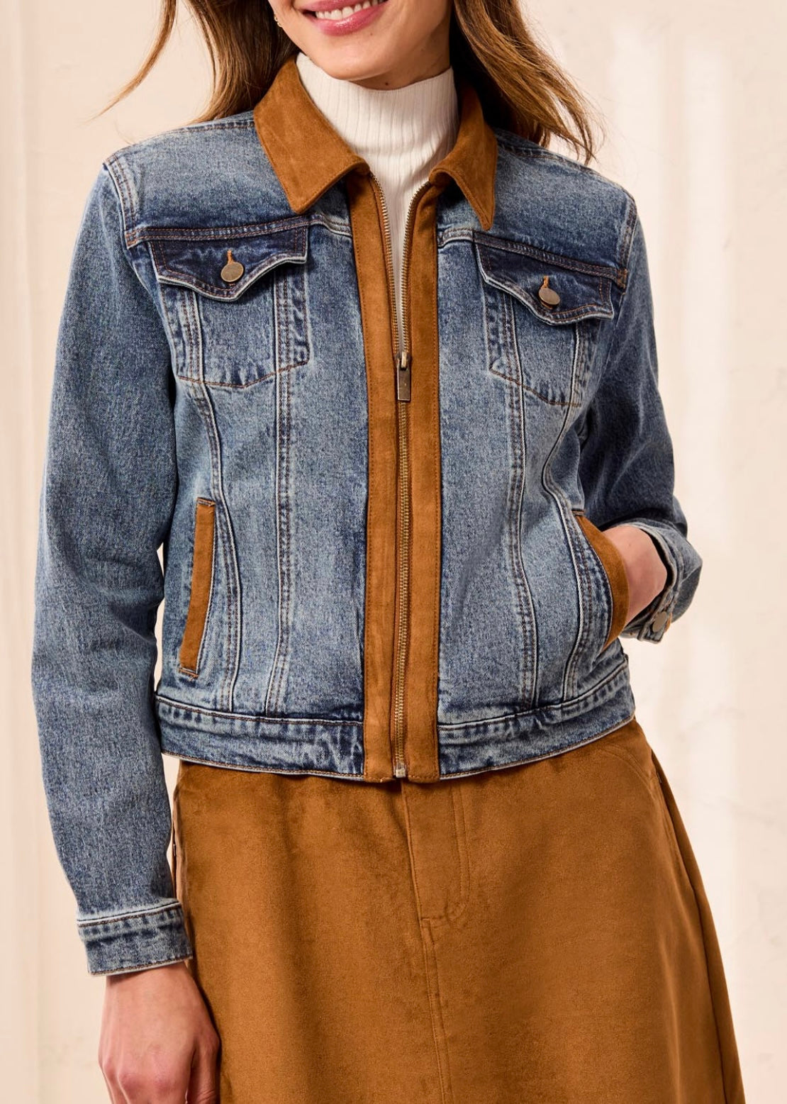 A woman wearing a cropped blue denim jacket with a brown collar and brown zipper trim, layered over a white turtleneck and brown skirt.