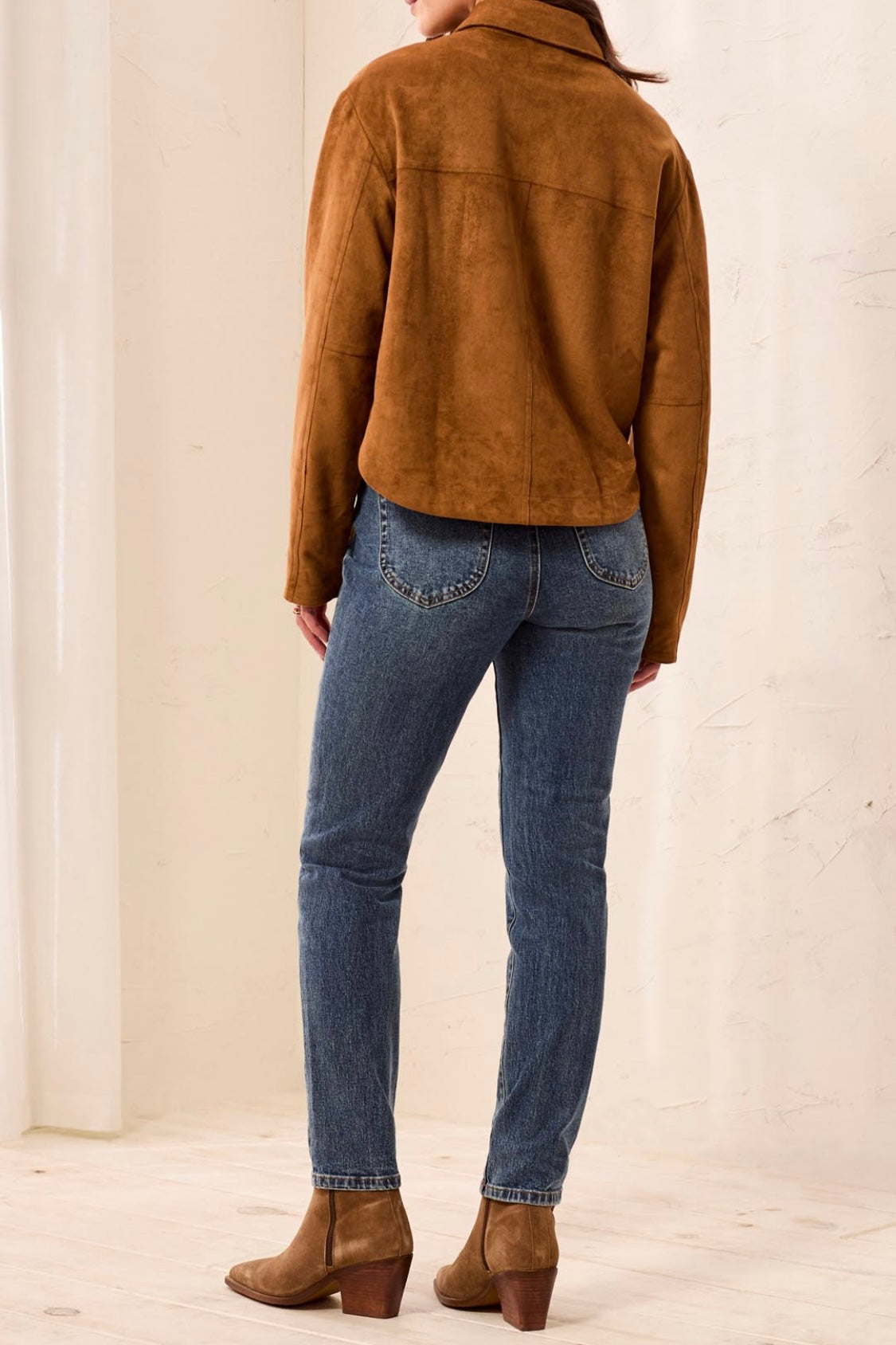 A model shown from the back wearing a brown suede jacket, blue skinny jeans, and brown suede ankle boots, standing against a light beige textured wall.