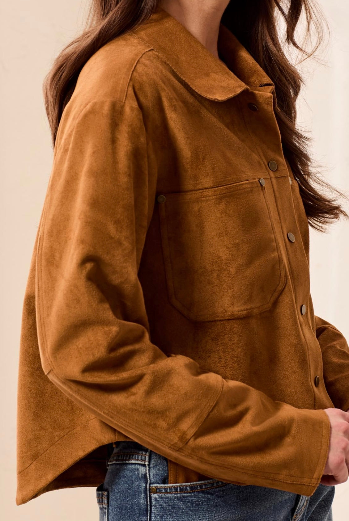 A close-up of a person wearing a cropped, long-sleeve brown suede or faux-suede jacket with a collar and front button placket, paired with blue jeans against a plain light beige background.