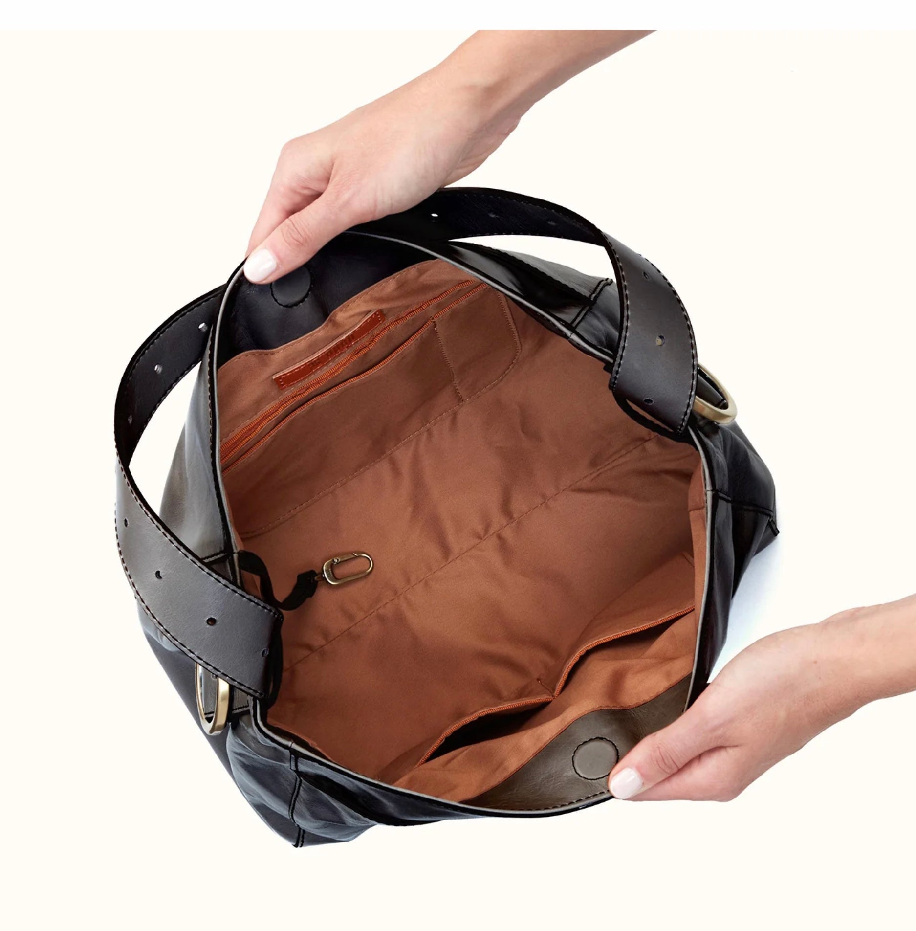 A black leather shoulder bag with a brown interior lining, shown open with hands holding it to display the spacious compartment and pockets.