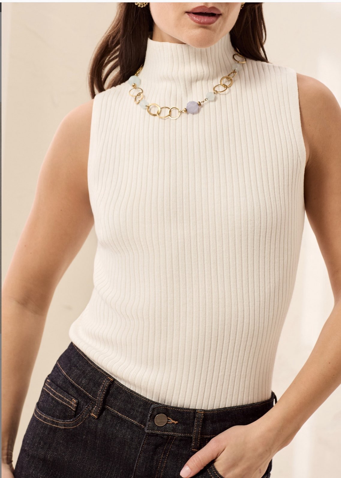 A woman wearing a ribbed sleeveless turtleneck top in off-white, paired with dark denim jeans and a gold chain necklace with a lavender stone.