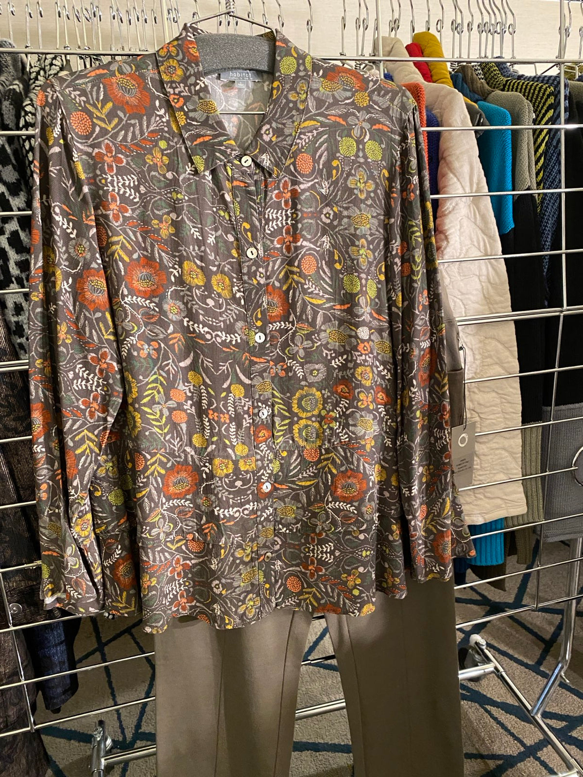 A folded short-sleeve button-up shirt with a busy multicolor floral and paisley print on a dark gray background, resting on a wire clothing rack.