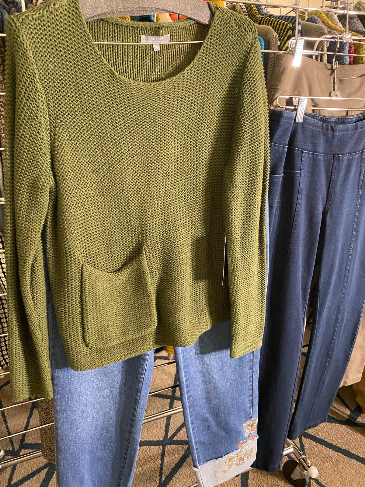An olive green knit sleeveless top with a large front patch pocket, laid flat over a pair of blue jeans on a clothing rack.