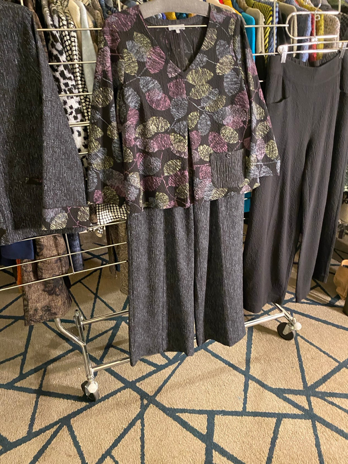 A clothing rack filled with folded and hanging garments, including dark gray knit tops and a floral-patterned top with purple, green, and gold tones, set against a patterned beige carpet.