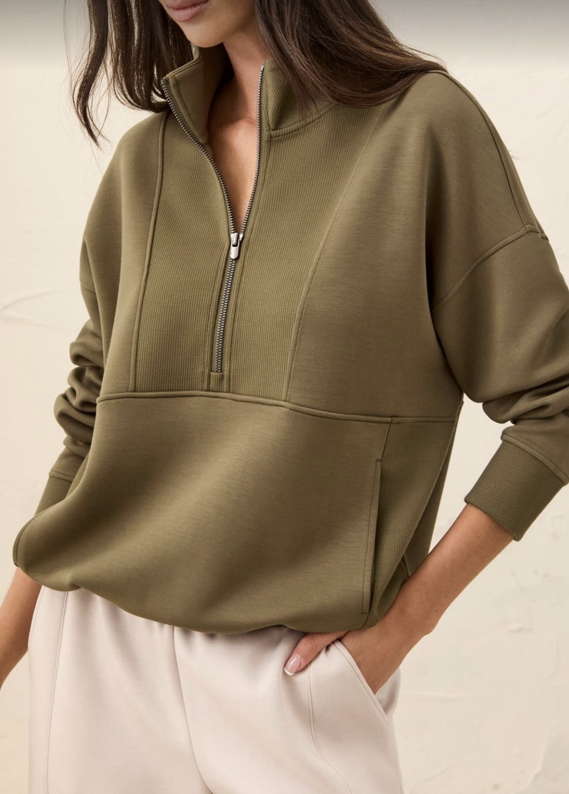 A woman wearing an olive green quarter-zip pullover sweatshirt with a stand collar and front pockets, paired with light cream pants, against a neutral beige background.