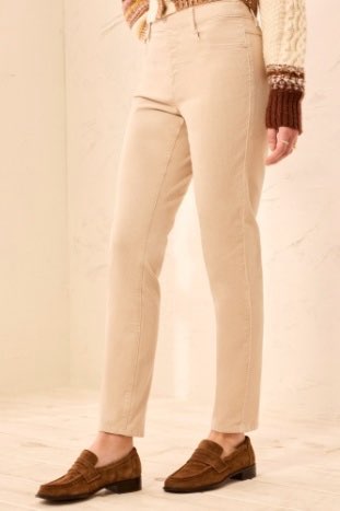 A person wearing straight-leg beige trousers, a cream knit sweater with brown cuffs, and brown suede loafers, shown from the waist down against a light beige wall and floor.