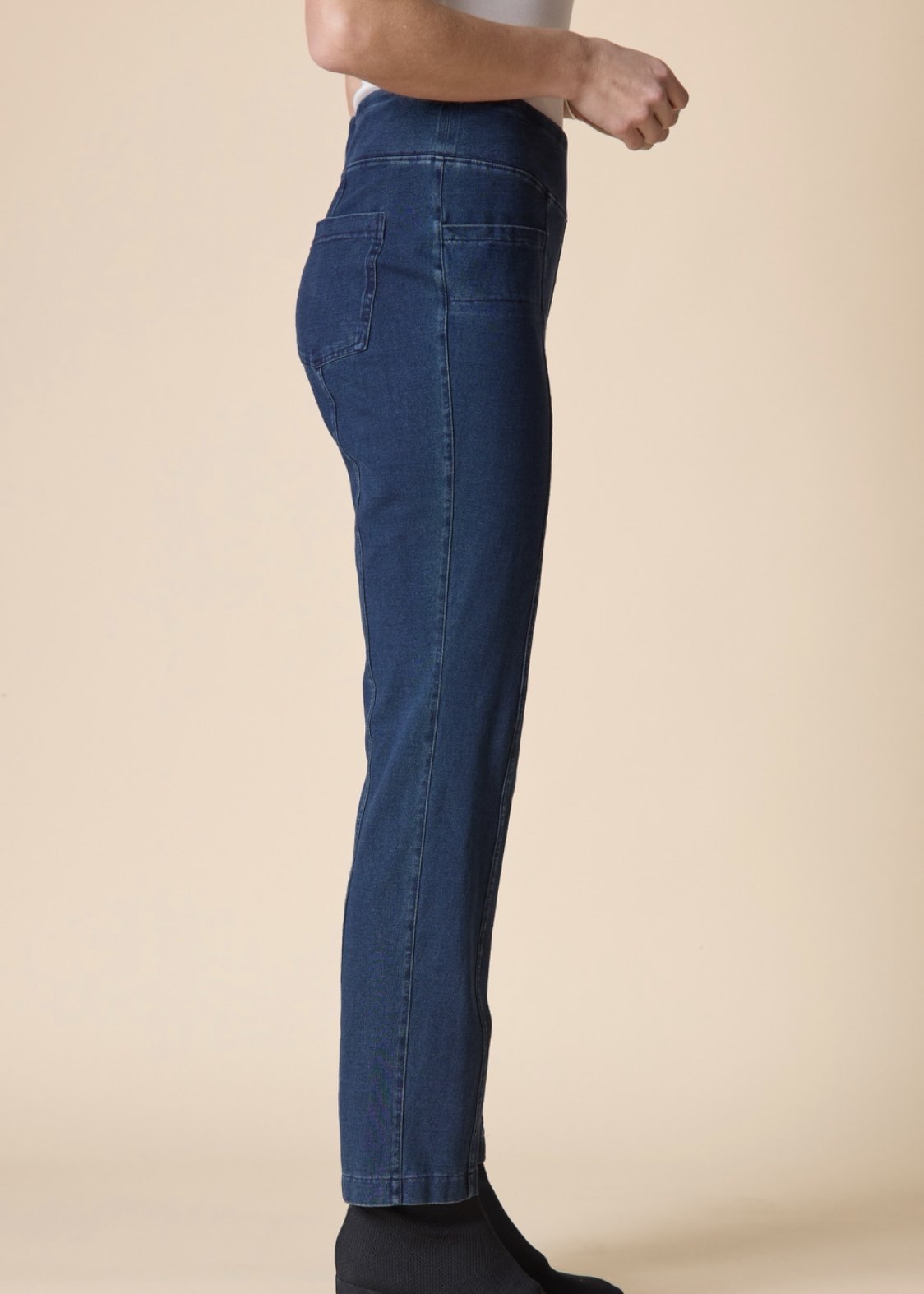 A side-profile view of a person wearing dark blue slim-fit jeans, shown from the waist down against a solid beige studio background.