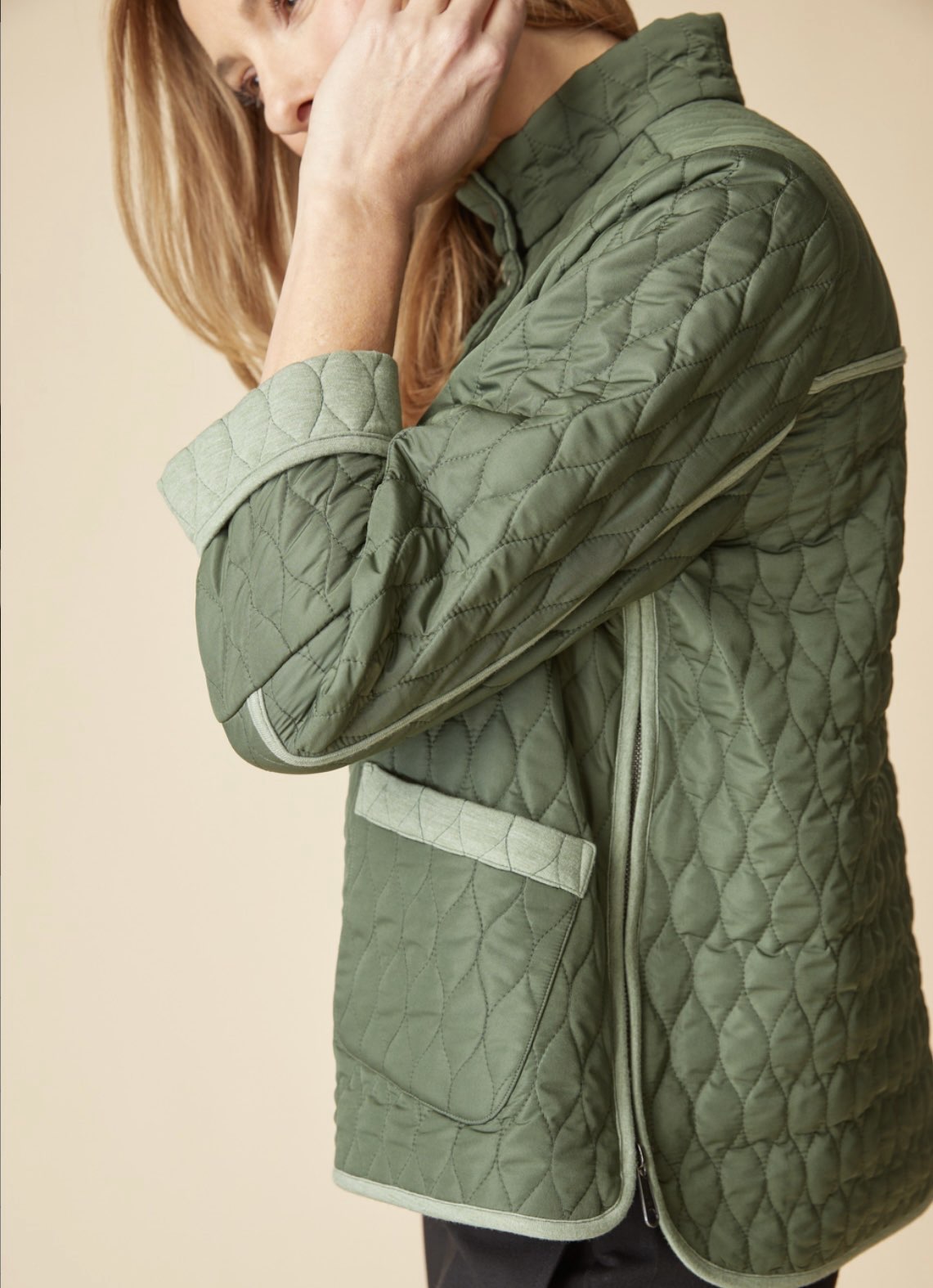A woman wearing a sage green quilted jacket with light beige trim, shown from a side-back angle against a solid beige background.