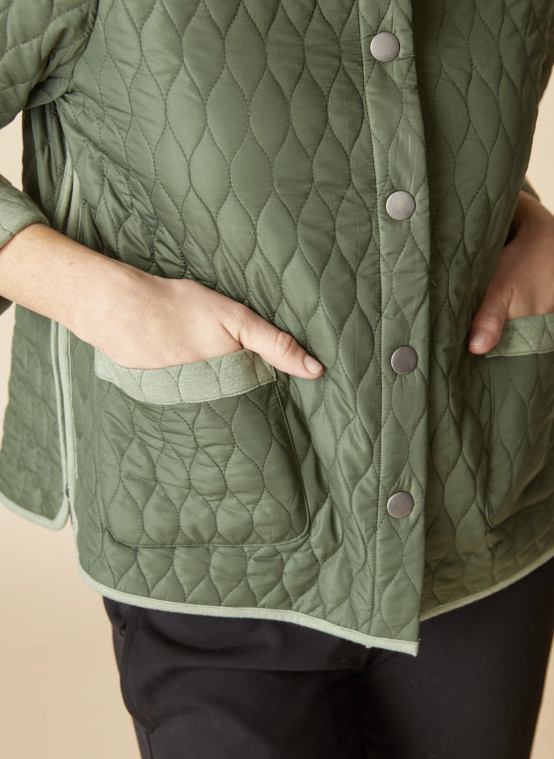 Close-up of a person wearing an olive green quilted jacket with a button front and patch pockets, paired with black pants against a plain beige background.