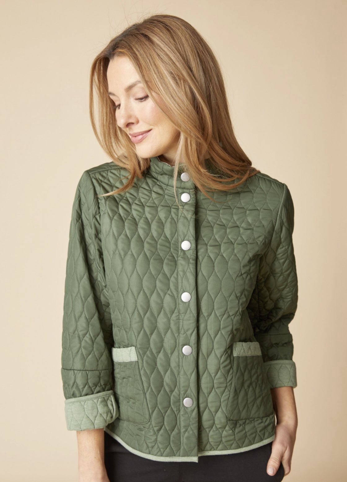 A woman wearing a quilted sage green jacket with a stand collar, button front, and ribbed cuffs, posed against a solid beige background.
