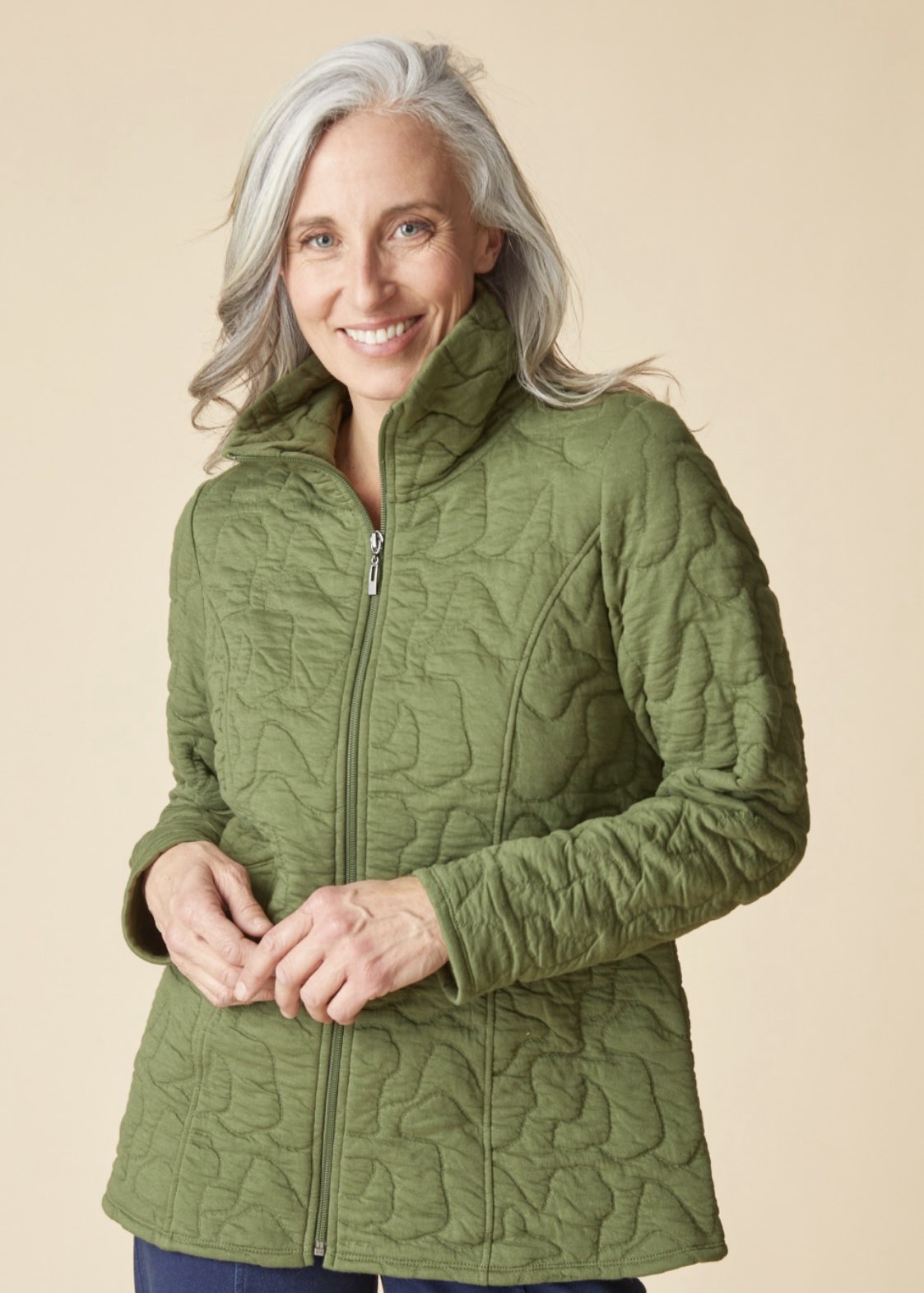 A smiling woman with gray hair wearing an olive green quilted zip-up jacket, standing against a solid beige background.