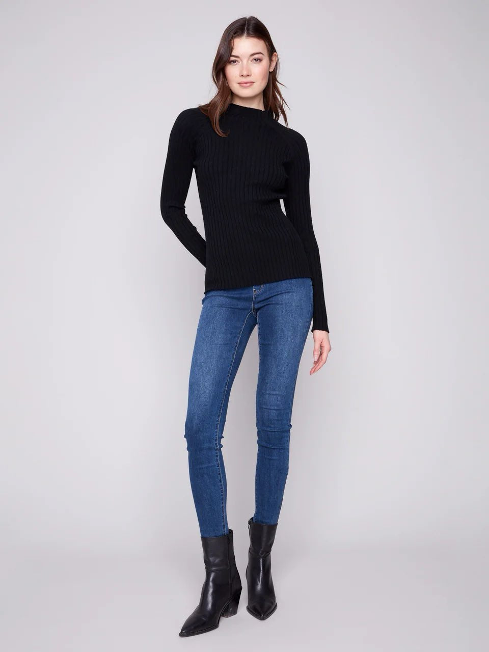 A woman modeling a black ribbed mock neck long-sleeve sweater, paired with blue skinny jeans and black ankle boots, against a light gray studio background.
