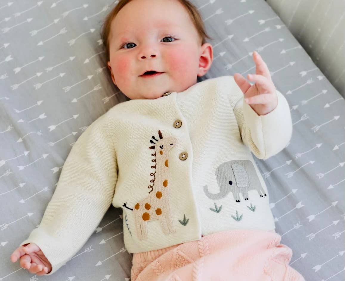 Baby wearing a cream-colored cardigan with animal designs on a crib with a patterned sheet.