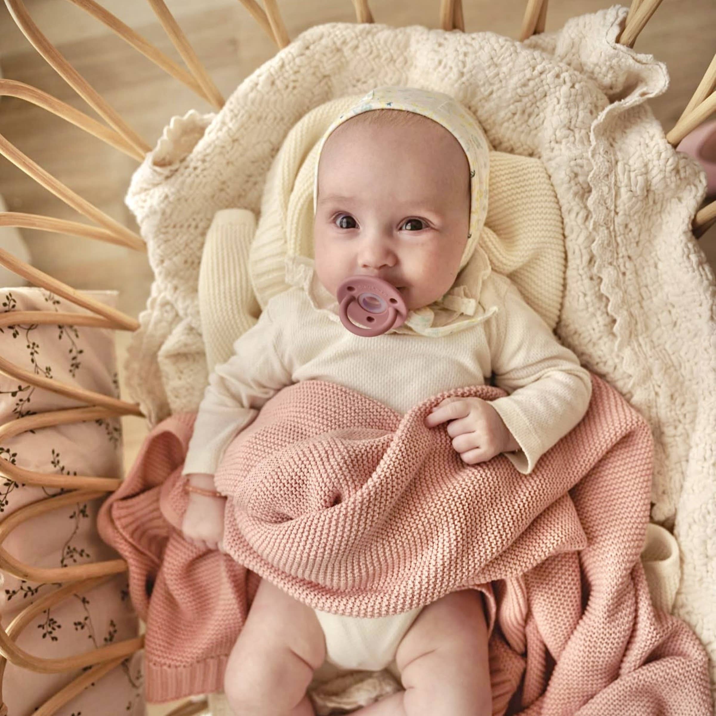 Baby in a crib with pink blanket and pacifier, surrounded by soft textures