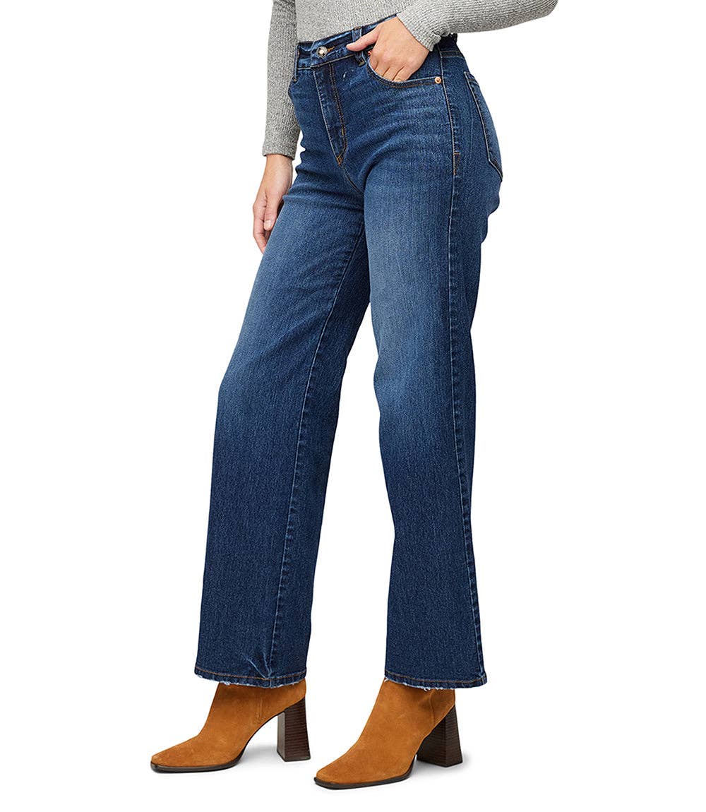 A model wearing a pair of high-waisted, wide-leg blue denim jeans with a straight-to-flare leg, styled with a gray long-sleeve top and brown suede block-heel ankle boots against a white background.