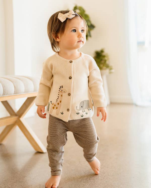 Baby in a beige cardigan and gray pants standing indoors.